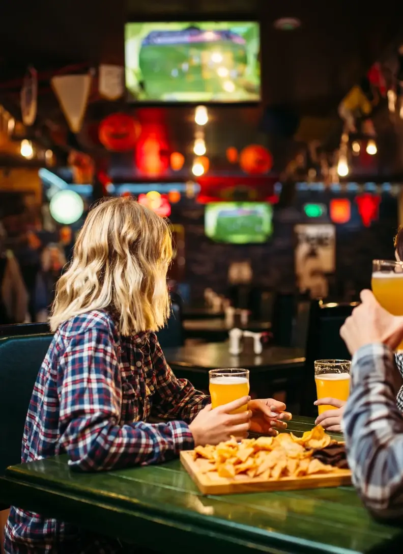 Two individuals are seated at a table in a dimly lit bar, sharing drinks and nachos with sports playing on screens in the background.