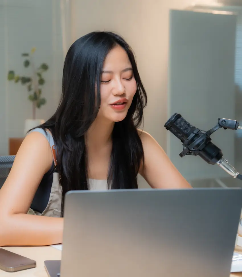 A young woman is recording voice announcements with a microphone in front of a laptop.