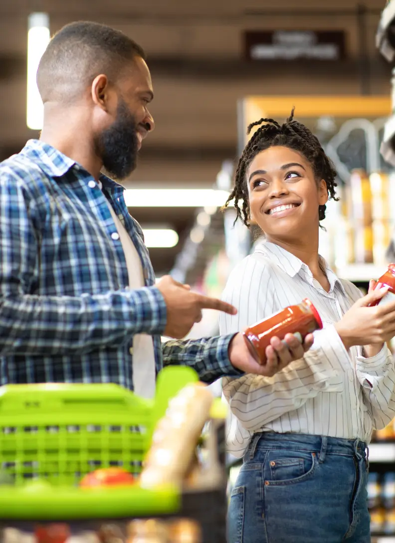 A young couple is grocery shopping, holding jars of pasta sauce.